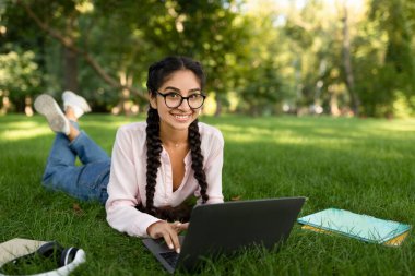 Açık havada okuyan mutlu Hintli kız öğrenci çimenlikte dizüstü bilgisayarla yatıyor, kameraya gülümsüyor, boş yer var.