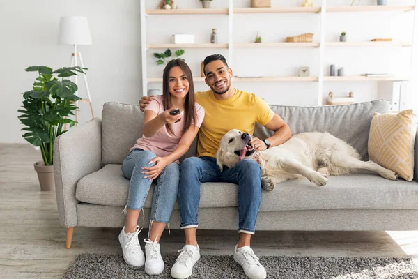 Jeune couple affectueux multiracial assis sur le canapé avec chien de compagnie et regarder la télévision à la maison — Photo