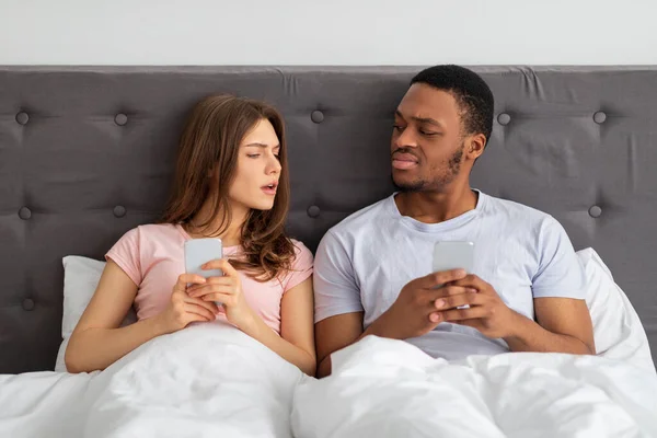 Jeune couple interracial avec des smartphones assis au lit, coincés dans des gadgets, se regardant avec suspicion — Photo