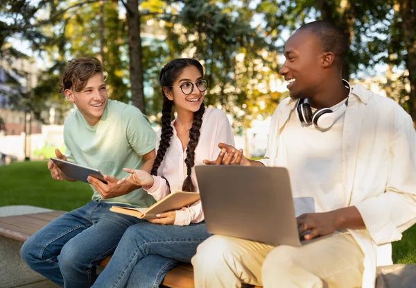 Estudiantes estilo. Tres amigos de la universidad aprendiendo juntos, usando computadoras portátiles y libros de lectura, sentados en el campus —  Fotos de Stock