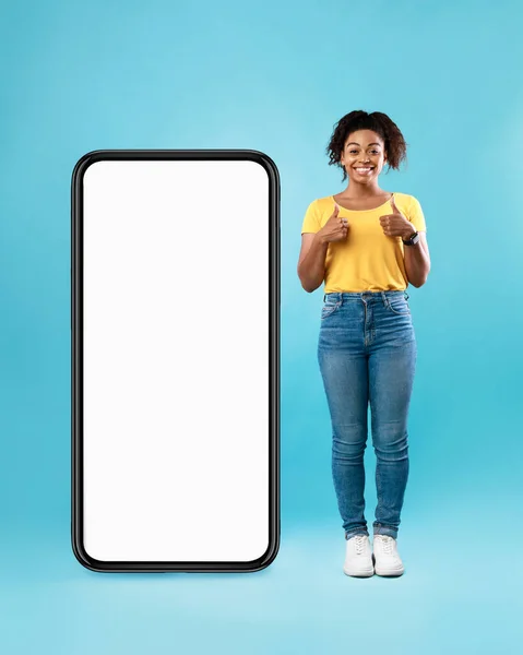 Усміхнена афро-леді демонструє гігантський смартфон з білим екраном і показує великі пальці вгору, макет для додатка або веб-сайту — стокове фото