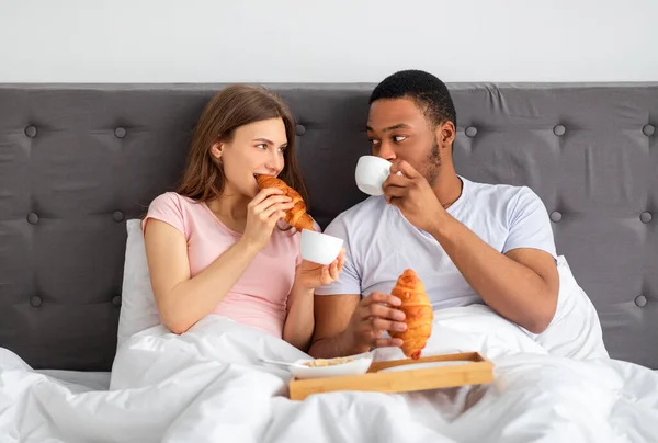 Romantica coppia multirazziale mangiare croissant freschi di crescita caffè, facendo colazione a letto a casa — Foto Stock