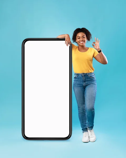 Pozitif siyah kadın büyük bir cep telefonunun yanında boş ekranla poz veriyor, iyi bir jest gösteriyor, web sitesi öneriyor, maket yapıyor. — Stok fotoğraf