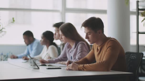 Educación fuera de línea. Vista lateral de jóvenes estudiantes multiétnicos diversos sentados en el aula y escribiendo notas — Vídeos de Stock