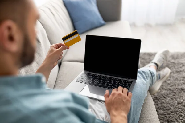 Αγνώριστος νεαρός που κάθεται στον καναπέ, χρησιμοποιώντας πιστωτική κάρτα για online αγορές σε laptop, χώρο για mockup ιστοσελίδα — Φωτογραφία Αρχείου