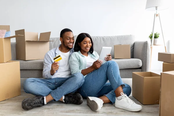 Entusiasmado casal negro escolhendo móveis on-line, comprando bens para seu novo apartamento, usando tablet digital e cartão de crédito — Fotografia de Stock