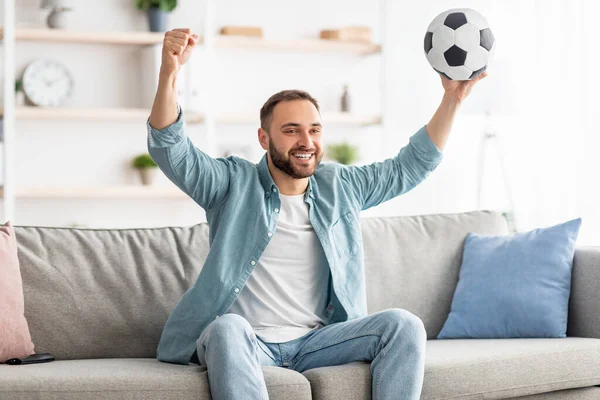 Aufgeregter junger Kerl schaut Fußball im Fernsehen, hebt Ball über den Kopf, feiert Tor oder Sieg auf der heimischen Couch — Stockfoto