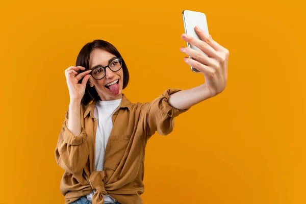 Speelse vrouw nemen selfie, tongzoenen op mobiele telefoon camera en aanraken bril, poseren over gele achtergrond — Stockfoto