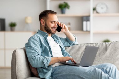 Akıllı telefondan konuşan çekici bir genç ve evdeki kanepede dizüstü bilgisayar kullanan.