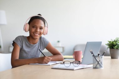 Mutlu Afro-Amerikan liseli kız kulaklıkları evde ve laptopuyla test ve sınava hazırlanıyor.