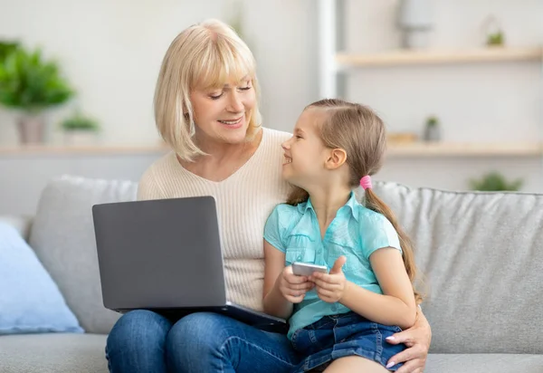 Happy mature woman and granddaughter using laptop and cell phone — Stockfoto