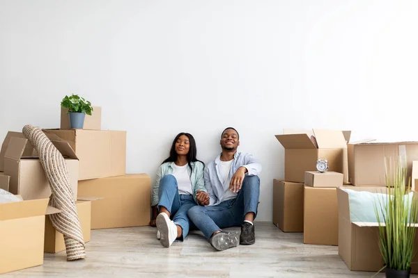 Felices esposos afroamericanos milenarios que se trasladan a un nuevo apartamento, sentados entre cajas de cartón, espacio vacío —  Fotos de Stock