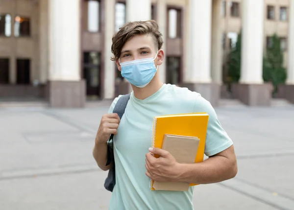 Diák fickó arcmaszkot visel, könyvekkel pózol, kint áll az egyetemi épület közelében, másolja a helyet — Stock Fotó