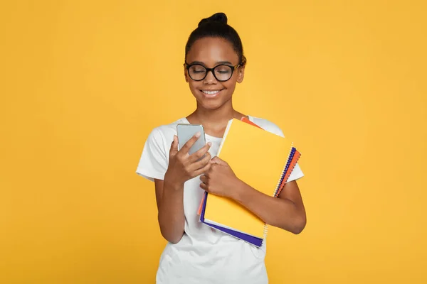 笑顔ティーンアフリカ系アメリカ人の女の子学生で眼鏡やカジュアルホールド本やスマートフォンでメッセージを読みます — ストック写真
