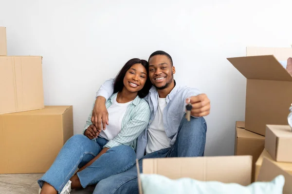 Propiedad de la casa. Amante pareja negra sosteniendo las llaves, sentado entre cajas de cartón y abrazos, espacio de copia — Foto de Stock