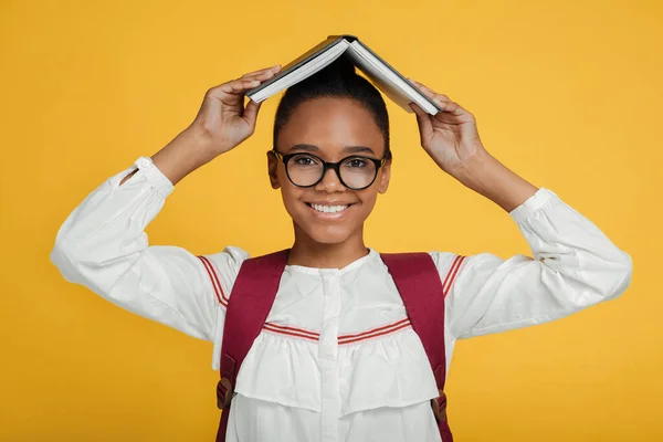 Smiling smart young african american female pupils in glasses with backpack holding book on her head — Stock Photo, Image