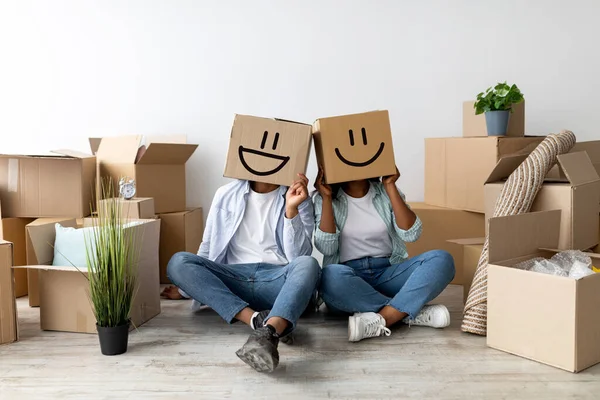 Verhuizen en vastgoed concept. Zwart paar met kartonnen dozen met smiley gezichten op het hoofd, zittend op de vloer — Stockfoto