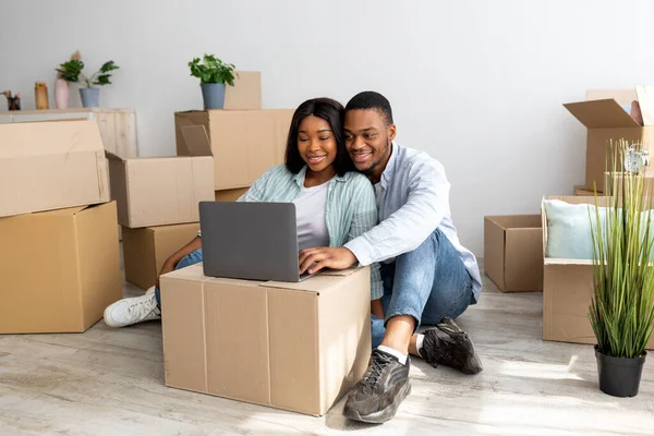 Felizes cônjuges afro-americanos tendo videochamada via laptop, sentados entre caixas de papelão em novo apartamento — Fotografia de Stock