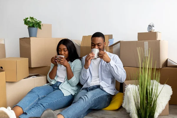 Χαλαρωμένο μαύρο ζευγάρι κάθεται ανάμεσα σε κουτιά από χαρτόνι και πίνοντας καφέ μετά τη μετάβαση σε νέο επίπεδο — Φωτογραφία Αρχείου