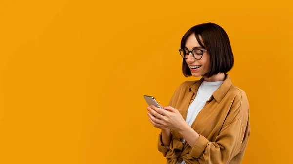 新的惊人的应用。快乐积极的年轻女士使用智能手机，站在黄色的背景上，全景 — 图库照片