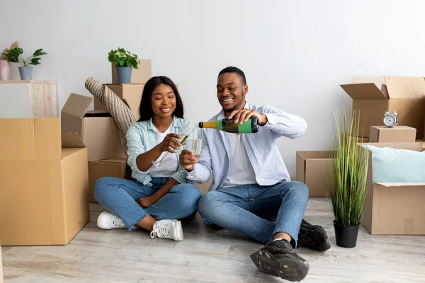 Amando a los cónyuges negros celebrando el día de mudanza con champán, sentados en el piso en un nuevo apartamento entre cajas de cartón — Foto de Stock