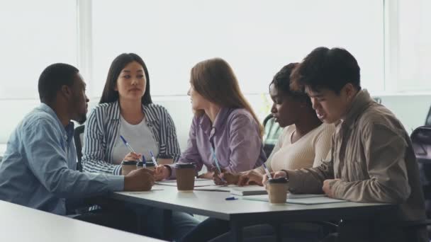 Grupo de estudiantes universitarios felices y diversos sentados en la biblioteca del campus, tomando café para llevar y haciendo deberes juntos — Vídeos de Stock