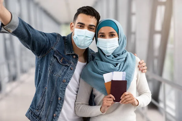 Счастливые исламские супруги в медицинских масках, делающие селфи в аэропорту — стоковое фото