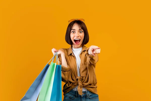 ¡Eh, tú! Mujer emocionada sosteniendo coloridas bolsas de papel de compras y apuntando a la cámara sobre fondo de estudio naranja — Foto de Stock