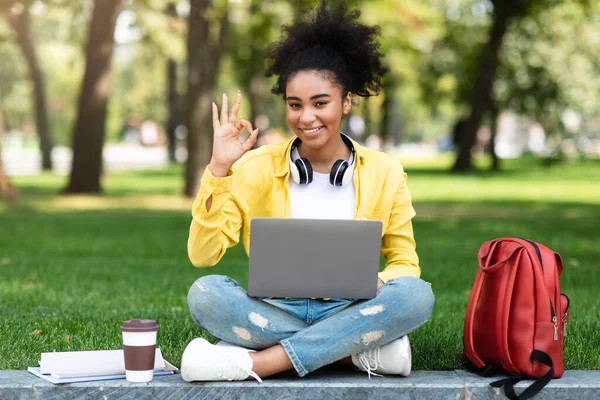 Nero studente ragazza utilizzando computer portatile gesturing ok simbolo seduta all'aperto — Foto Stock
