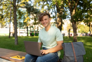 E-öğrenme. İnternette konferansa katılan heyecanlı erkek öğrenci dışarıda parkta bankta oturuyor.