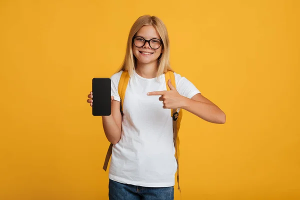 Happy female pupil in white t-shirt and glasses with backpack shows finger at smartphone with empty screen — Stock fotografie