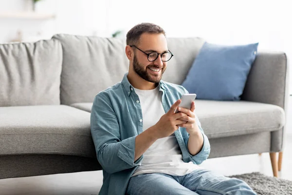 Chico joven feliz en gafas usando el teléfono celular, sentado en el suelo en la sala de estar, trabajando o comunicándose en línea desde casa — Foto de Stock