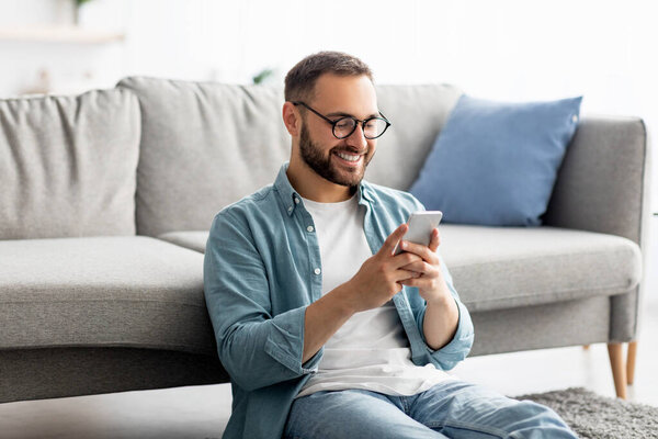 Happy young guy in glasses using cellphone, sitting on floor in living room, working or communicating online from home