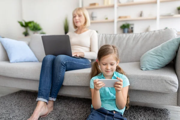 Mature woman and granddaughter using laptop and cell phone — Stock Photo, Image