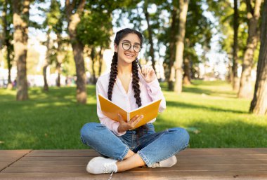 Not defterine dışarıda yazan mutlu Hintli kız öğrenci kampüs parkında bankta oturuyor. Bayan parkta boş vakit geçirirken ders çalışıyor. Modern öğrenme ve bilgi, sınava hazırlanma