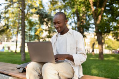 Laptop kullanan ve internetten öğrenen Afro-Amerikan erkek öğrenci dışarıda üniversite kampüs parkında ödev yapıyor. Bilgisayarda yazan, uzaktan çalışan bir adam.