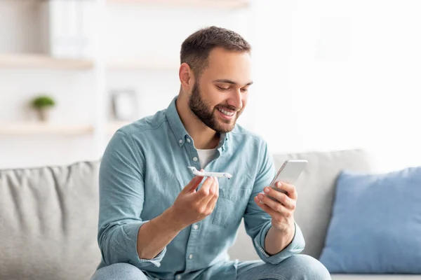 Retrato de un joven alegre sosteniendo un avión de juguete y usando un teléfono celular para comprar boletos o reservar vacaciones en línea desde casa — Foto de Stock