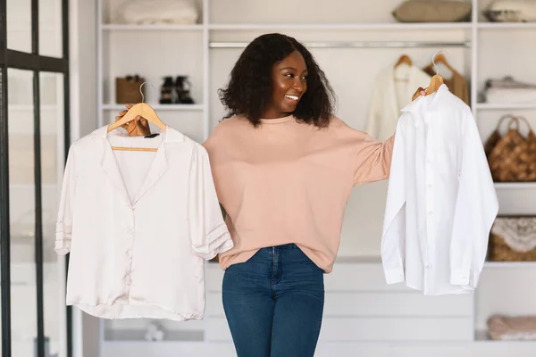 Mujer africana feliz comprando y eligiendo ropa sosteniendo camisas en interiores — Foto de Stock