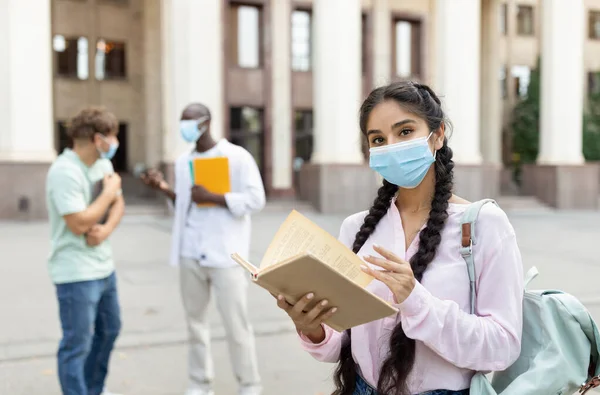 女学生戴着面罩，摆姿势看书，看着相机，站在大学大楼附近 — 图库照片