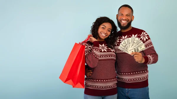 幸せなアフリカ系アメリカ人配偶者でクリスマスセーター保持ショッピングバッグとお金の束以上青背景 — ストック写真
