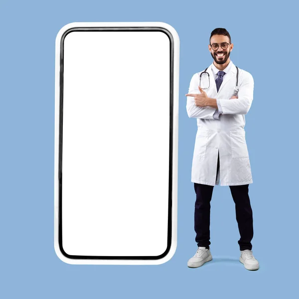 Árabe masculino médico posando perto Grande telefone tela, fundo azul — Fotografia de Stock