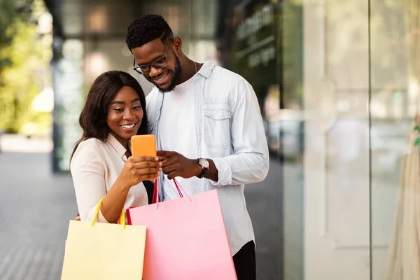 Happy black couple using phone holding shopping bags — Stock Photo, Image