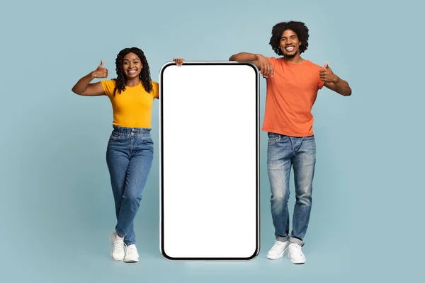 快乐的非洲裔美国千禧夫妇 在一个有着空白屏幕和大拇指的巨大的新手机旁随意地站着 推荐最新的移动应用程序 蓝色工作室背景 — 图库照片