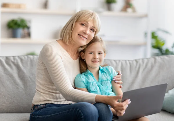 笑着的老奶奶和可爱的小孙女一起在笔记本电脑上看卡通片 快乐的老奶奶在玩游戏 坐在沙发上拥抱 看着相机 — 图库照片
