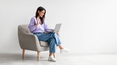 Gülümseyen genç bir kadın arkadaşlarıyla ya da ailesiyle video görüşmesi yapıyor, dizüstü bilgisayar kullanıyor, webcam 'e el sallıyor, rahat koltukta oturuyor.