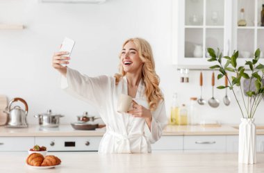 Avrupa 'dan gülümseyen, bornozlu, sarışın, kahve içen, kahvaltıda akıllı telefondan selfie çeken bir kadın.