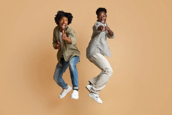 Dos alegres chicos negros apuntando a la cámara mientras saltan sobre el fondo beige —  Fotos de Stock