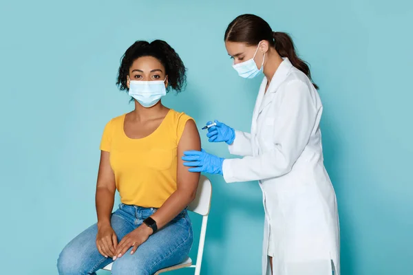 Giovane medico che fa l'iniezione di vaccino anti-covid alla giovane donna afroamericana in maschera, sfondo blu studio — Foto Stock