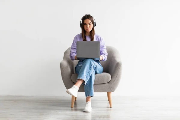Ung vit kvinna som har videosamtal, använder bärbar dator, sitter i fåtölj, bär trådlösa hörlurar — Stockfoto