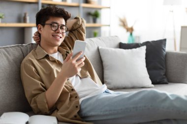 Gözlüklü gülümseyen Asyalı adam evde akıllı telefon kullanıyor.